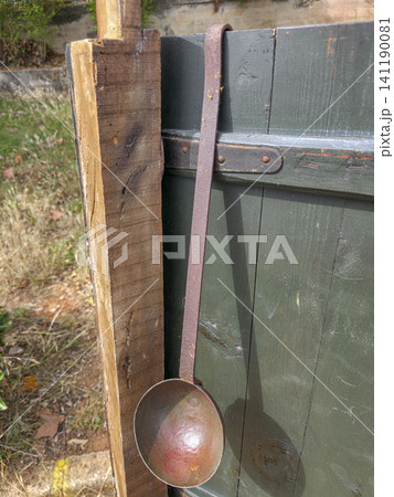 A rustic metal ladle is hanging on a dark green wooden fence panel. The setting is outdoors with some grass and dirt visible. The ladle shows signs of wear and age. A rustic metal ladle is hanging on a dark green wooden fence panel. The setting is outdoors with some grass and dirt visible. The ladle shows signs of wear and age. 141190081