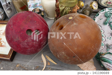 XALO, SPAIN - JUNE 20, 2026.Two scratched bowling balls are on a table at a flea market. One ball is red and the other is brown amber. Various vintage items surround them. 141190089