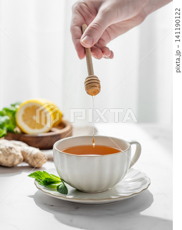 Hand holding a honey stick over a glass cup of tea with lemon, ginger,  mint on a marble background 141190122