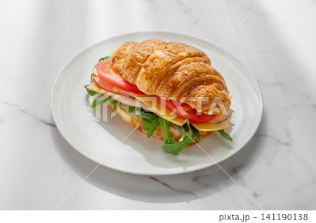 A golden croissant sandwich with ham, cheese, tomato, and arugula on a marble background A golden croissant sandwich with ham, cheese, tomato, and arugula on a marble background 141190138