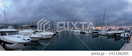 Luxury marina shows many sailing yachts and motorboats lined up as twilight sets in. Dark clouds fill the sky, creating a dramatic atmosphere over the water. Luxury marina shows many sailing yachts and motorboats lined up as twilight sets in. Dark clouds fill the sky, creating a dramatic atmosphere over the water. 141190265