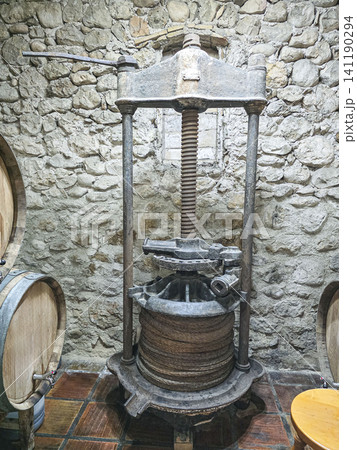 An ancient grape press is located in a rustic wine cellar with stone walls. Wooden barrels are nearby, holding wine. This scene shows the process of winemaking from the past. 141190294