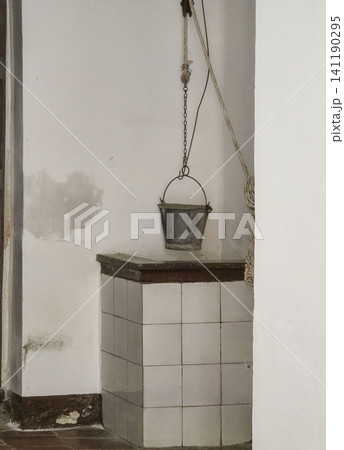 Inside an apartment of an old traditional Spanish house, a historic water well stands with a metal bucket hanging from the ceiling, showcasing simple architecture. Inside an apartment of an old traditional Spanish house, a historic water well stands with a metal bucket hanging from the ceiling, showcasing simple architecture. 141190295