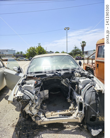 A white car sits in a junkyard with its engine and front bumper removed. The scene is under a clear blue sky with other vehicles in the background. It shows the state of vehicle recycling. A white car sits in a junkyard with its engine and front bumper removed. The scene is under a clear blue sky with other vehicles in the background. It shows the state of vehicle recycling. 141190300