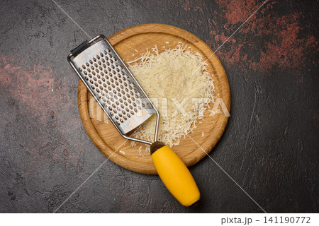 A pile of grated cheese and a metal grater on a round wooden board, dark textured background A pile of grated cheese and a metal grater on a round wooden board, dark textured background 141190772