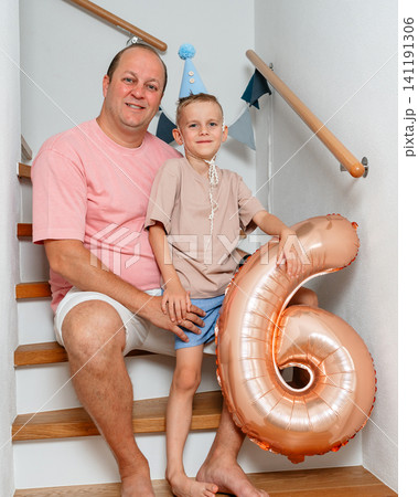 father and son sitting on wooden stairs with pink balloon number six and textile party hat, celebrating a birthday in an indoor setting, front view, family portraits, birthday celebrations. father and son sitting on wooden stairs with pink balloon number six and textile party hat, celebrating a birthday in an indoor setting, front view, family portraits, birthday celebrations. 141191306