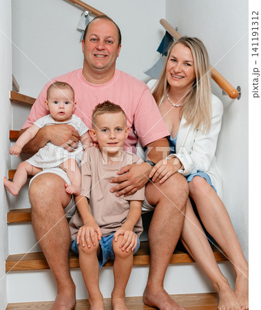 A family of four, including a father, mother, son, and baby girl, is sitting together on a wooden staircase adorned with pink and white textiles, creating a relaxed and natural atmosphere. A family of four, including a father, mother, son, and baby girl, is sitting together on a wooden staircase adorned with pink and white textiles, creating a relaxed and natural atmosphere. 141191312