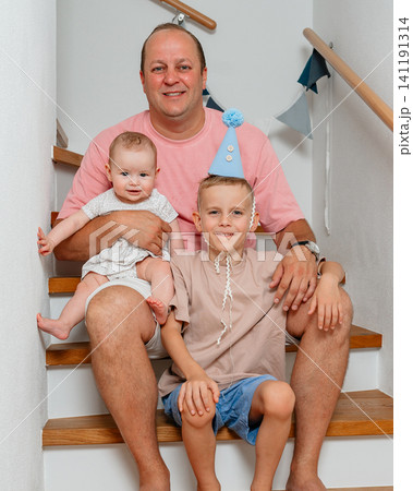 a joyful family of three sitting together on a wooden staircase in their home, surrounded by white walls and a blue party hat, capturing the essence of family portraits, parent-child bond. 141191314