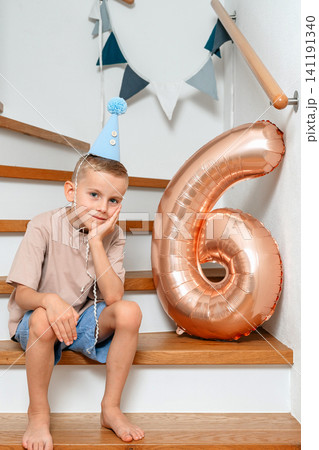 a boy sits on stairs with a party hat and number '6' balloon, celebrating his birthday in an indoor staircase adorned with decorative flags and wooden railings, captured from a frontal view. a boy sits on stairs with a party hat and number '6' balloon, celebrating his birthday in an indoor staircase adorned with decorative flags and wooden railings, captured from a frontal view. 141191340