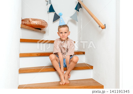 Boy sitting on stairs, in indoor staircase with white walls and wooden handrail, with staircase, wooden handrail, party hat, balloon, with wood, plastic texture, from a frontal view perspective. Boy sitting on stairs, in indoor staircase with white walls and wooden handrail, with staircase, wooden handrail, party hat, balloon, with wood, plastic texture, from a frontal view perspective. 141191345