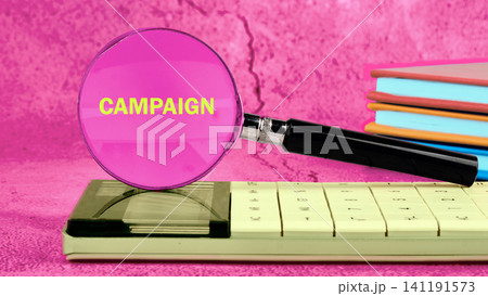 A magnifying glass highlights the word campaign on a calculator that sits on a colorful stack of notebooks on a desk in a bright setting 141191573