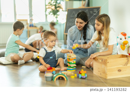 Children playing in living room of kindergarten, babysitter support and participate in game. Children playing in living room of kindergarten, babysitter support and participate in game. 141193282