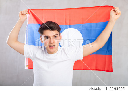 Reliant positive young man holds big national flag of Laos with confident smile showing teeth. Reliant positive young man holds big national flag of Laos with confident smile showing teeth. 141193665