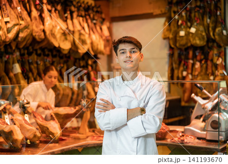 Portrait of experienced butcher on background of rack with hanging various jamon 141193670
