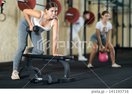 Athletic young woman pumping her biceps with a dumbbell while leaning on bench 141193716