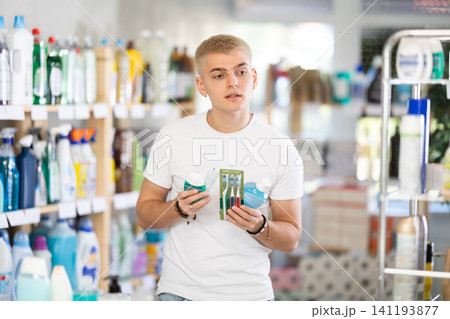 Positive young man choosing toothpaste and toothbrush, dental kit in store 141193877