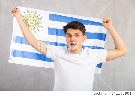 Reliant positive young man holds big national flag of Uruguay with confident smile showing teeth Reliant positive young man holds big national flag of Uruguay with confident smile showing teeth 141193901
