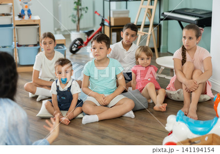 Group of children is sitting on floor, listening to teacher in kindergarten Group of children is sitting on floor, listening to teacher in kindergarten 141194154