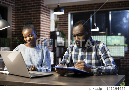 Coworkers discuss stock figures in nighttime office during a office setting moment involving african american, real time and tech savvy Coworkers discuss stock figures in nighttime office during a office setting moment involving african american, real time and tech savvy 141194433