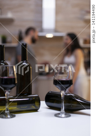 Empty bottles and wine glasses stand on a kitchen counter while an intoxicated couple sits behind them, showing alcohol abuse and unhealthy home drinking. Empty bottles and wine glasses stand on a kitchen counter while an intoxicated couple sits behind them, showing alcohol abuse and unhealthy home drinking. 141194490