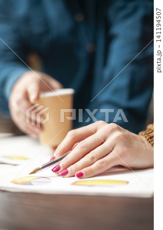 Woman points with a pen at financial documents near coffee on a table, emphasizing paperwork review, selective focus and company research in an office task. 141194507