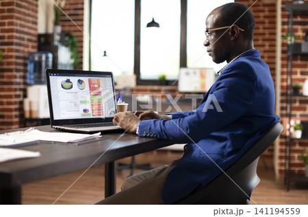 Black businessman studies financial analysis on a computer screen at his desk, reviewing market strategy and company data in a startup office. Black businessman studies financial analysis on a computer screen at his desk, reviewing market strategy and company data in a startup office. 141194559