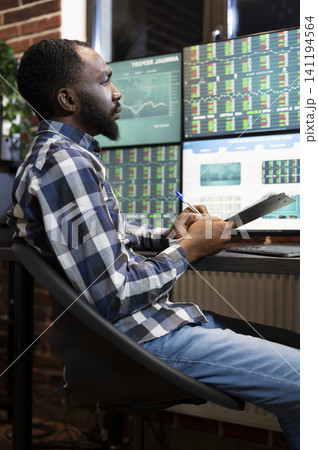 Stockbroker writes notes while studying forex market trends on multiple monitors at night, reviewing financial data and stock charts, with chart analysis and trading workspace context. Stockbroker writes notes while studying forex market trends on multiple monitors at night, reviewing financial data and stock charts, with chart analysis and trading workspace context. 141194564