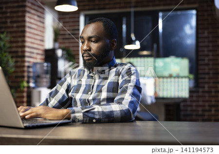 African American businessman works late at a desk, typing on a laptop and researching forex market data for stock investment decisions, with chart analysis and trading workspace context. African American businessman works late at a desk, typing on a laptop and researching forex market data for stock investment decisions, with chart analysis and trading workspace context. 141194573