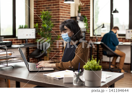Businesswoman wearing a face mask reviews financial charts on a laptop at a neat desk in a brick wall office while checking startup company data. 141194644