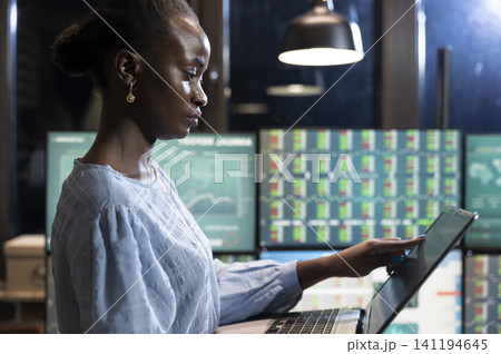 African American stockbroker stands at her workstation with laptop and multiple monitors while reviewing financial reports and investment data at night. African American stockbroker stands at her workstation with laptop and multiple monitors while reviewing financial reports and investment data at night. 141194645