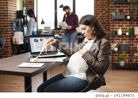 Pregnant businesswoman reviewing research reports showing pregnant businesswoman sitting and reviewing research data at office desk with laptop nearby. 141194655