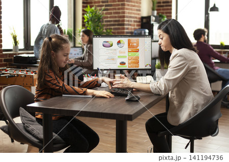 Playful daughter interrupting her mom as she tackles complex financial data on computer monitor. Caucasian little girl sitting and playing around on table, disturbing tired mother at work. Playful daughter interrupting her mom as she tackles complex financial data on computer monitor. Caucasian little girl sitting and playing around on table, disturbing tired mother at work. 141194736