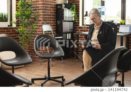Elderly therapist sits alone with clipboard and glasses in a brick wall room, reviewing patient notes before a counselling session with clients. Elderly therapist sits alone with clipboard and glasses in a brick wall room, reviewing patient notes before a counselling session with clients. 141194742
