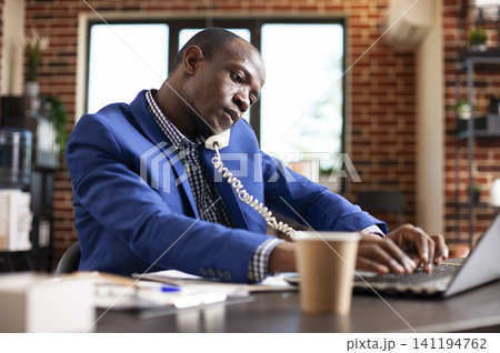 Office manager handling call and laptop in brick wall office while working through documents, digital reports and company planning tasks. Office manager handling call and laptop in brick wall office while working through documents, digital reports and company planning tasks. 141194762
