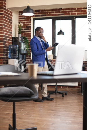 African american manager using digital device to research market insights in brick wall room, with black male analyst standing with tablet and contemplating company project development in startup. African american manager using digital device to research market insights in brick wall room, with black male analyst standing with tablet and contemplating company project development in startup. 141194810
