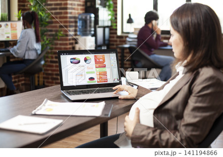 Pregnant entrepreneur reviews market analysis on a laptop at an office desk, planning financial strategy in a startup workplace for realistic business use and specific office workflow. 141194866