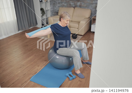 Senior woman sitting on balance ball training with resistance band. Old person training at home sport healthy lifestyle, elderly fitness exercise workout in apartment, activity. Senior woman sitting on balance ball training with resistance band. Old person training at home sport healthy lifestyle, elderly fitness exercise workout in apartment, activity. 141194976