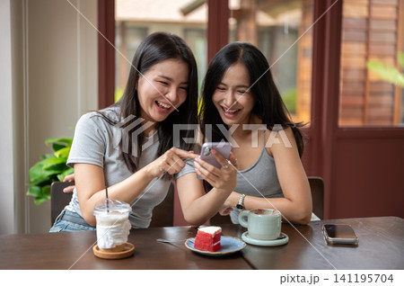 Asian women friends browsing internet on smartphone together over coffee and cake at table in cafe. Asian women friends browsing internet on smartphone together over coffee and cake at table in cafe. 141195704