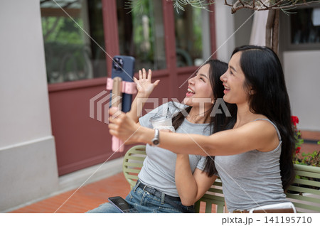 Asian women friends enjoy taking an outdoor selfie together in a bright and relaxing cafe courtyard. Asian women friends enjoy taking an outdoor selfie together in a bright and relaxing cafe courtyard. 141195710