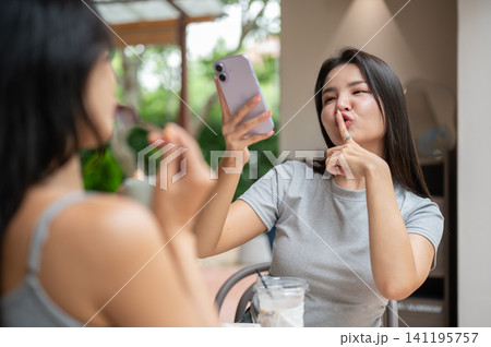 Funny Asian woman holding smartphone up high while making a hushing finger gesture at a cafe. Funny Asian woman holding smartphone up high while making a hushing finger gesture at a cafe. 141195757