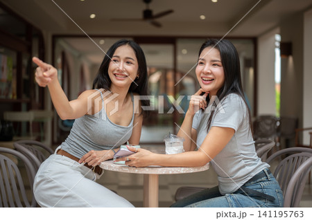 oyful Asian woman pointing away while sitting at a coffee shop table with her friend. oyful Asian woman pointing away while sitting at a coffee shop table with her friend. 141195763