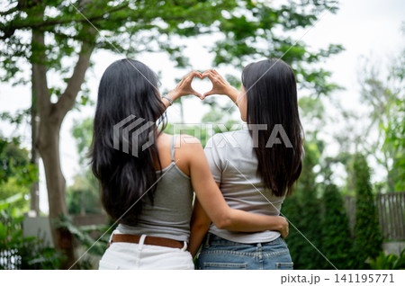 Rear view of two Asian women hugging each other and making a heart shape hand gesture in the park. 141195771
