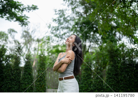 Relaxed Asian woman hugging herself with eyes closed on a green lawn. Relaxed Asian woman hugging herself with eyes closed on a green lawn. 141195780
