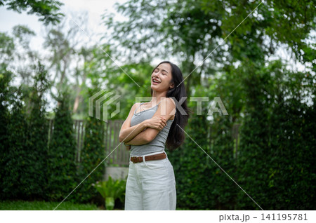 Charming Asian woman hugging herself and laughing cheerfully in a green backyard. Charming Asian woman hugging herself and laughing cheerfully in a green backyard. 141195781
