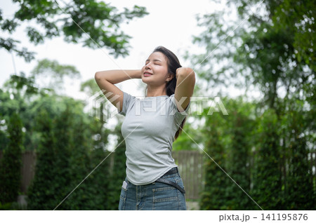 Relaxed Asian woman holding hands behind head and closing eyes in a garden. Relaxed Asian woman holding hands behind head and closing eyes in a garden. 141195876