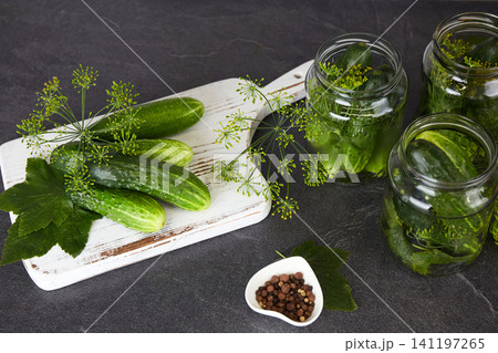 Cucumbers in glass jars and on a cutting board, dill, garlic, and spices on a white wooden background. Cucumbers in glass jars and on a cutting board, dill, garlic, and spices on a white wooden background. 141197265
