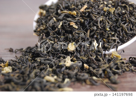 Close up of dried jasmine tea leaves spilling from a white ceramic spoon onto a wooden surface. Close up of dried jasmine tea leaves spilling from a white ceramic spoon onto a wooden surface. 141197698