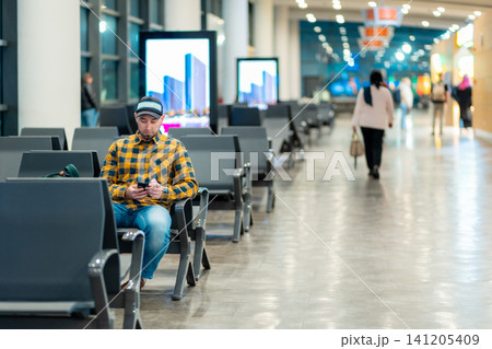 Wide shot of adult Caucasian man in cap and jacket using smartphone in airport waiting area sitting in chair. Copy space. Concept of travel and trip Wide shot of adult Caucasian man in cap and jacket using smartphone in airport waiting area sitting in chair. Copy space. Concept of travel and trip 141205409