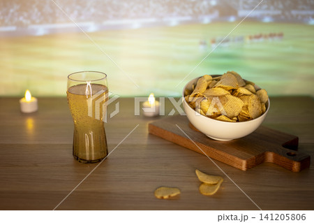 A glass of beer and potato chips snack on wooden table during soccer match, highlighting comforting food for big football events. A glass of beer and potato chips snack on wooden table during soccer match, highlighting comforting food for big football events. 141205806