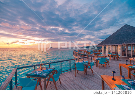 Overwater restaurant deck with set tables at dramatic sunset in tropical resort. Wooden terrace with blue chairs facing colorful evening sky over calm sea. Luxury alfresco dining venue for hospitality 141205810